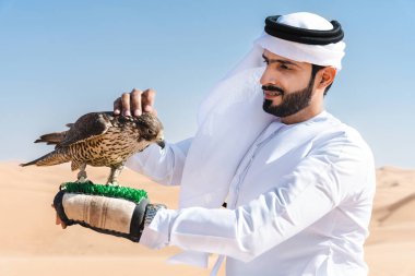 Ortadoğulu bir adam çölde geleneksel emirati Arap kandurası giyiyor ve Dubai 'deki kum tepelerinde Arap Müslüman bir şahin kuşu tutuyor.
