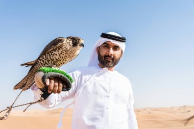 Ortadoğulu bir adam çölde geleneksel emirati Arap kandurası giyiyor ve Dubai 'deki kum tepelerinde Arap Müslüman bir şahin kuşu tutuyor.