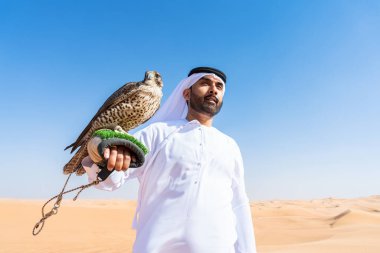 Ortadoğulu bir adam çölde geleneksel emirati Arap kandurası giyiyor ve Dubai 'deki kum tepelerinde Arap Müslüman bir şahin kuşu tutuyor.