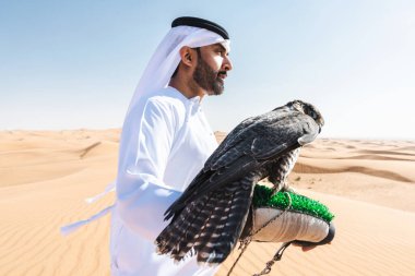 Ortadoğulu bir adam çölde geleneksel emirati Arap kandurası giyiyor ve Dubai 'deki kum tepelerinde Arap Müslüman bir şahin kuşu tutuyor.