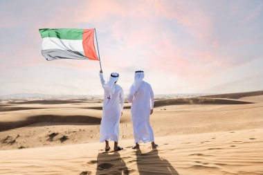 Two middle-eastern men wearing traditional emirati arab kandura bonding in the desert and holiding the UAE flag to celebrate national day - Arabian muslim friends meeting at the sand dunes in Dubai clipart