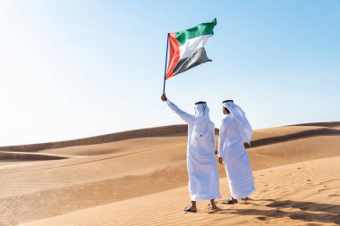 Geleneksel emirati Arap kandurası giyen iki orta doğulu adam çölde kaynaşıyor ve ulusal bayrağı kutluyorlar - Arap Müslümanlar Dubai 'de kum tepelerinde buluşuyorlar