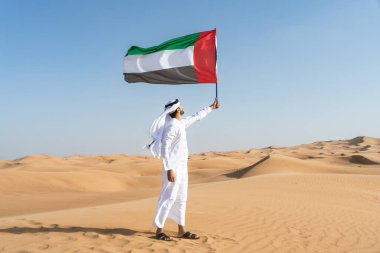 Çölde geleneksel emirati Arap kandurası giyen ve elinde BAE bayrağı tutan Ortadoğulu bir adam Dubai 'deki kum tepelerinde ulusal vatanseverliği kutlayan Müslüman bir Arap bayrağı taşıyor.