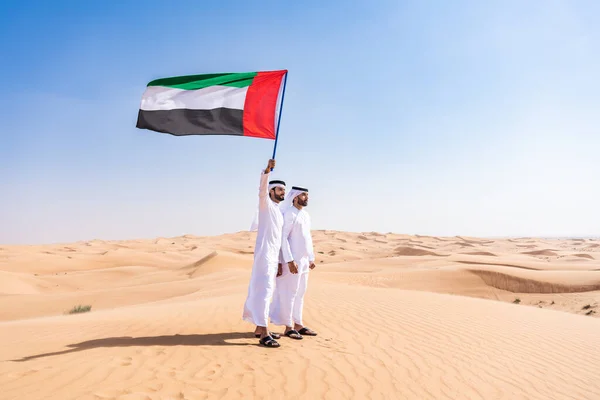 Two middle-eastern men wearing traditional emirati arab kandura bonding in the desert and holding emirati flag -  Arabian muslim friends meeting at the sand dunes in Dubai