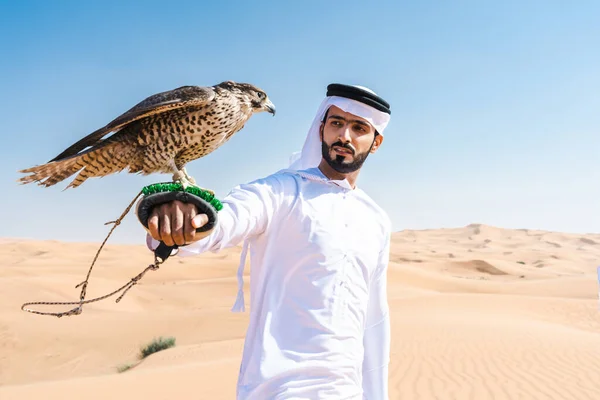 砂漠で伝統的なエミレーツ アラブ カンドゥラを身に着けている中東の男と鷹の鳥を保持 ドバイの砂丘でアラブのイスラム教徒の大人の人 — ストック写真