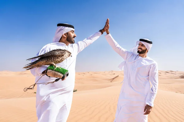 Two middle-eastern men wearing traditional emirati arab kandura bonding in the desert and holding a falcon bird - Arabian muslim friends meeting at the sand dunes in Dubai