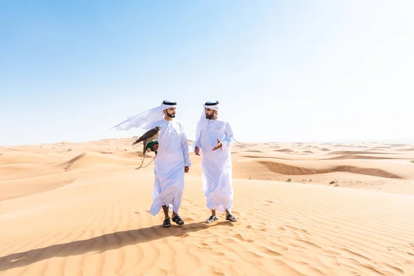 砂漠で伝統的なエミレーツ アラブ カンドゥラのボンディングを身に着けている中東の2人の男性と鷹の鳥を保持している ドバイの砂丘でアラビア人のイスラム教徒の友人と会う — ストック写真