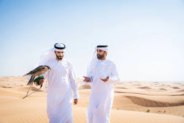Two middle-eastern men wearing traditional emirati arab kandura bonding in the desert and holding a falcon bird - Arabian muslim friends meeting at the sand dunes in Dubai