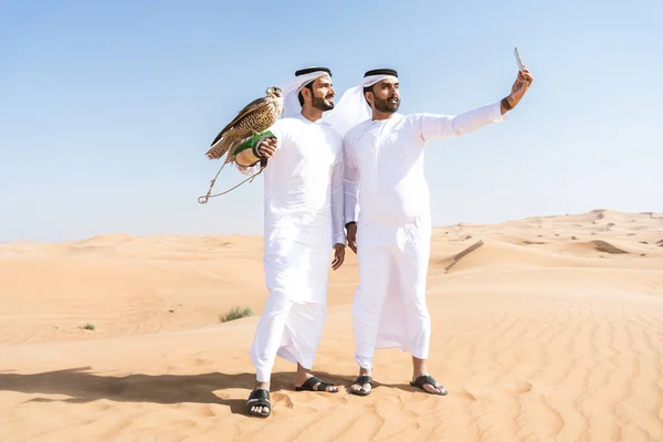 砂漠で伝統的なエミレーツ アラブ カンドゥラのボンディングを身に着けている中東の2人の男性と鷹の鳥を保持している ドバイの砂丘でアラビア人のイスラム教徒の友人と会う — ストック写真