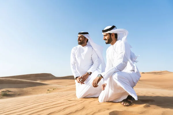 Two middle-eastern men wearing traditional emirati arab kandura bonding in the desert - Arabian muslim friends meeting at the sand dunes in Dubai