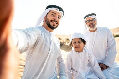 Dubai çölünde beyaz kandura kıyafeti içinde safari yapan üç kuşak aile. Büyükbaba, oğul ve torun doğada birlikte vakit geçiriyorlar..