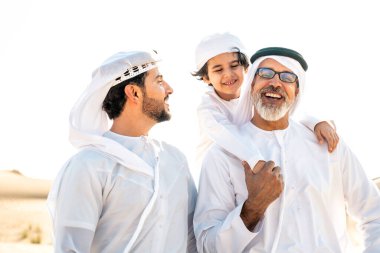 Dubai çölünde beyaz kandura kıyafeti içinde safari yapan üç kuşak aile. Büyükbaba, oğul ve torun doğada birlikte vakit geçiriyorlar..
