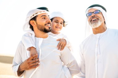 Dubai çölünde beyaz kandura kıyafeti içinde safari yapan üç kuşak aile. Büyükbaba, oğul ve torun doğada birlikte vakit geçiriyorlar..