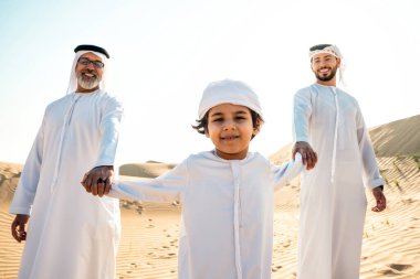 Dubai çölünde beyaz kandura kıyafeti içinde safari yapan üç kuşak aile. Büyükbaba, oğul ve torun doğada birlikte vakit geçiriyorlar..