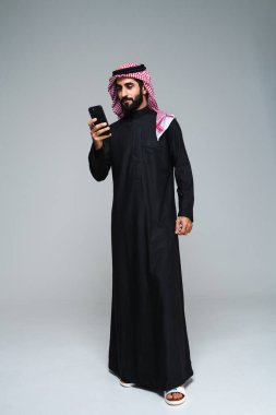 Stüdyoda geleneksel Suudi kıyafetleri olan yakışıklı Arap orta doğulu adam - Arap Müslüman yetişkin iş adamı beyaz arka planda izole edilmiş thwab portresi takıyor.