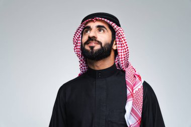 Stüdyoda geleneksel Suudi kıyafetleriyle yakışıklı bir Arap orta doğulu adam - Arap Müslüman yetişkin erkek işadamı, gri arka planda izole edilmiş twab portresi giyiyor