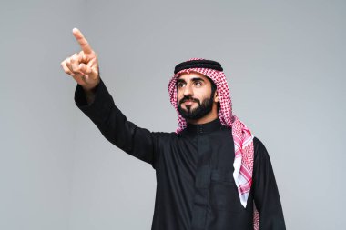 Stüdyoda geleneksel Suudi kıyafetleriyle yakışıklı bir Arap orta doğulu adam - Arap Müslüman yetişkin erkek işadamı, gri arka planda izole edilmiş twab portresi giyiyor
