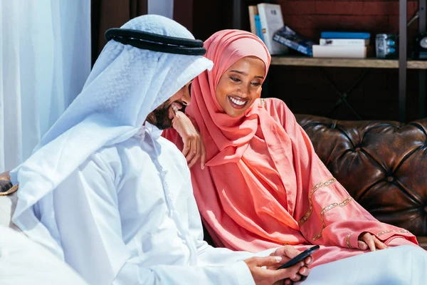 Happy middle-eastern couple wearing traditional arab clothing at home - Married arabian husband and wife bonding together in the apartment, concepts about relationship, domestic life and emirati lifestyle