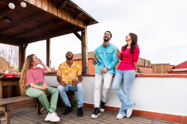 Çok ırklı bir grup mutlu genç arkadaş evlerinin çatısında barbekü partisi veriyorlar - çok ırklı neşeli genç insanlar teras balkonunda eğleniyor ve kaynaşıyorlar şehir manzarası ile, yiyip içiyorlar.