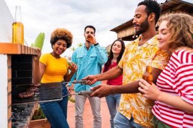Çok ırklı bir grup mutlu genç arkadaş evlerinin çatısında barbekü partisi veriyorlar - çok ırklı neşeli genç insanlar teras balkonunda eğleniyor ve kaynaşıyorlar şehir manzarası ile, yiyip içiyorlar.