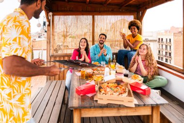 Çok ırklı bir grup mutlu genç arkadaş evlerinin çatısında barbekü partisi veriyorlar - çok ırklı neşeli genç insanlar teras balkonunda eğleniyor ve kaynaşıyorlar şehir manzarası ile, yiyip içiyorlar.