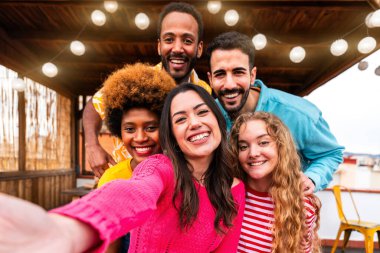 Çok ırklı bir grup mutlu genç arkadaş evlerinin çatısında barbekü partisi veriyorlar - çok ırklı neşeli genç insanlar teras balkonunda eğleniyor ve kaynaşıyorlar şehir manzaralı, yiyip içiyorlar. Komik yüzlerin yakın plan portresi.