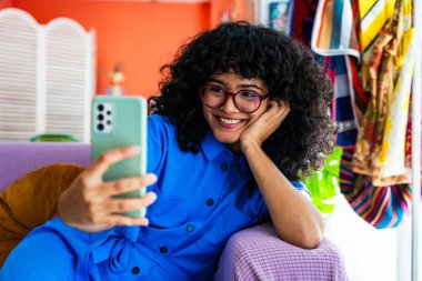 Evde kıvırcık saçlı güzel bir İspanyol kadın, sosyal medyada paylaşmak için akıllı telefonuyla kanepede selfie çekiyor.