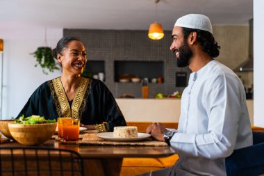 Emirati kıyafetli mutlu genç Arap çift evde öğle yemeği yiyorlar. Geleneksel Arap kıyafetli orta doğulu erkek ve kadın ramazan üstüne sağlıklı yiyecekler yiyorlar. 