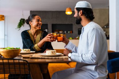 Emirati kıyafetli mutlu genç Arap çift evde öğle yemeği yiyorlar. Geleneksel Arap kıyafetli orta doğulu erkek ve kadın ramazan üstüne sağlıklı yiyecekler yiyorlar. 