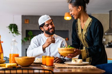 Emirati kıyafetli mutlu genç Arap çift evde öğle yemeği yiyorlar. Geleneksel Arap kıyafetli orta doğulu erkek ve kadın ramazan üstüne sağlıklı yiyecekler yiyorlar. 