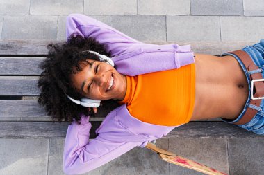 Açık havada kıvırcık saçlı güzel siyahi bir kadın modern havalı kadın şık şehir kıyafetleri giyiyor şehirde kaykayla geziyor.