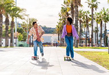 Açık havada kaykay yapan çok ırklı genç bir çift - iki arkadaş şehir bölgelerinde kayak yaparken eğleniyor, gençlik, çeşitlilik, yeşil traksiyonlar ve hareketlilik kavramları