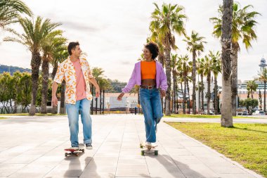 Açık havada kaykay yapan çok ırklı genç bir çift - iki arkadaş şehir bölgelerinde kayak yaparken eğleniyor, gençlik, çeşitlilik, yeşil traksiyonlar ve hareketlilik kavramları