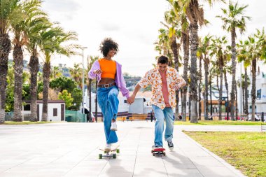 Açık havada kaykay yapan çok ırklı genç bir çift - iki arkadaş şehir bölgelerinde kayak yaparken eğleniyor, gençlik, çeşitlilik, yeşil traksiyonlar ve hareketlilik kavramları