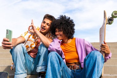 Açık havada kaykay yapan çok ırklı genç bir çift - iki arkadaş şehir bölgelerinde kayak yaparken eğleniyor, gençlik, çeşitlilik, yeşil traksiyonlar ve hareketlilik kavramları