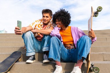 Açık havada kaykay yapan çok ırklı genç bir çift - iki arkadaş şehir bölgelerinde kayak yaparken eğleniyor, gençlik, çeşitlilik, yeşil traksiyonlar ve hareketlilik kavramları