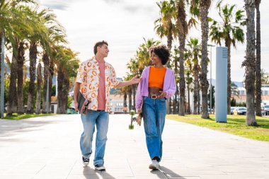 Açık havada kaykay yapan çok ırklı genç bir çift - iki arkadaş şehir bölgelerinde kayak yaparken eğleniyor, gençlik, çeşitlilik, yeşil traksiyonlar ve hareketlilik kavramları