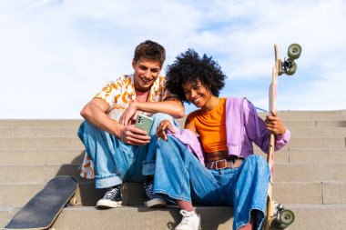 Açık havada kaykay yapan çok ırklı genç bir çift - iki arkadaş şehir bölgelerinde kayak yaparken eğleniyor, gençlik, çeşitlilik, yeşil traksiyonlar ve hareketlilik kavramları