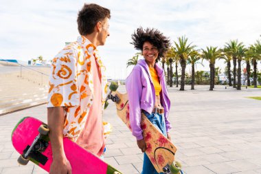 Açık havada kaykay yapan çok ırklı genç bir çift - iki arkadaş şehir bölgelerinde kayak yaparken eğleniyor, gençlik, çeşitlilik, yeşil traksiyonlar ve hareketlilik kavramları