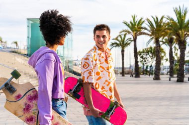 Açık havada kaykay yapan çok ırklı genç bir çift - iki arkadaş şehir bölgelerinde kayak yaparken eğleniyor, gençlik, çeşitlilik, yeşil traksiyonlar ve hareketlilik kavramları