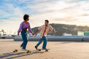 Açık havada kaykay yapan çok ırklı genç bir çift - iki arkadaş şehir bölgelerinde kayak yaparken eğleniyor, gençlik, çeşitlilik, yeşil traksiyonlar ve hareketlilik kavramları