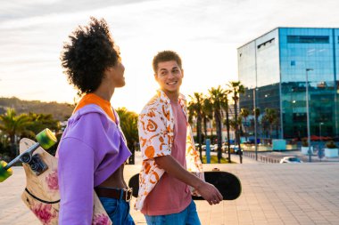 Açık havada kaykay yapan çok ırklı genç bir çift - iki arkadaş şehir bölgelerinde kayak yaparken eğleniyor, gençlik, çeşitlilik, yeşil traksiyonlar ve hareketlilik kavramları