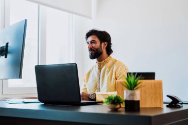 Ofisteki Pakistanlı iş adamı. Ofisinde geleneksel kıyafetleriyle çalışan genç bir iş adamı. Birden fazla aygıt ekranından alım satımlar kontrol ediliyor ve teknoloji kullanılıyor.
