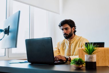 Ofisteki Pakistanlı iş adamı. Ofisinde geleneksel kıyafetleriyle çalışan genç bir iş adamı. Birden fazla aygıt ekranından alım satımlar kontrol ediliyor ve teknoloji kullanılıyor.