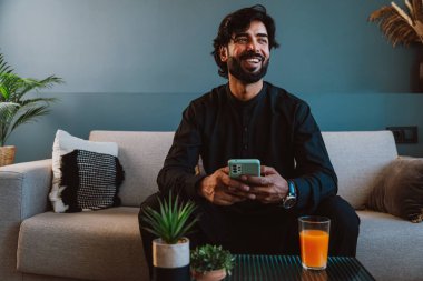 Geleneksel kıyafetli Pakistanlı genç adam koltukta akıllı telefon kullanıyor..