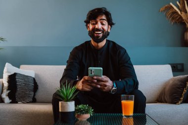 Geleneksel kıyafetli Pakistanlı genç adam koltukta akıllı telefon kullanıyor..