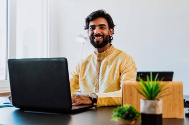 Ofisteki Pakistanlı iş adamı. Ofisinde geleneksel kıyafetleriyle çalışan genç bir iş adamı. Birden fazla aygıt ekranından alım satımlar kontrol ediliyor ve teknoloji kullanılıyor.