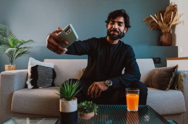 Geleneksel kıyafetli Pakistanlı genç adam koltukta akıllı telefon kullanarak selfie çekiyor..