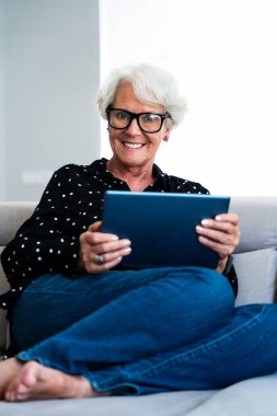 Güzel, yaşlı, gri saçlı, tablet kullanan yaşlı bir kadın. Genç, yaşlı bir büyükanne evde kaliteli zaman geçiriyor.