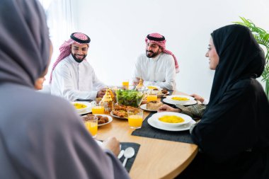 Suudi Arabistan kıyafeti giyen mutlu Arap ailesi akşam yemeği sırasında evde birlikte vakit geçiriyorlar - Orta Doğu Suudi aileleri ve çocukları Ramazan 'ı gün be gün kutluyorlar.